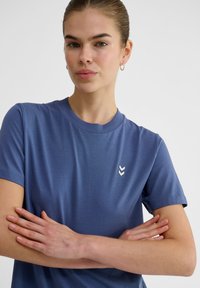 Blå kortärmad t-shirt i mjukt tyg med rund halsringning och en vit chevronlogotyp på vänster bröstområde. Enkelt och figursytt design.