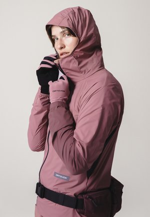 Person wearing a mauve hooded jacket with black and pink gloves, adjusting the zipper near the face, against a plain light background.