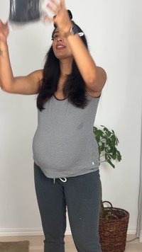 Noseča ženska v črtasti majici in sivih hlačah dviguje prozoren plastični vsebnik nad glavo v notranjosti, blizu lončnice.