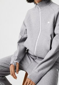 Šedá mikina na zip z měkké látky s vysokým límcem, bílým zipem a logem na hrudi. Kombinována se šedými kalhotami.