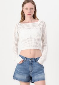 Hvit, cropsydd genser i åpen strikk med sommerfuglmønster på brystet, kombinert med blå denimshorts med knappefeste og lommer.