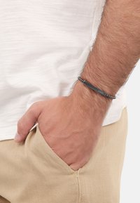 Bracciale in metallo nero con design a barra piatta, caratterizzato da accenti di perline e una superficie liscia, indossato su un polso con una camicia di colore chiaro.