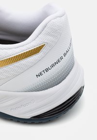 Hvide atletiske sko med tekstureret mesh overdel, guld- og sorte accenter, FlyteFoam branding og gradientdesign på hælen.