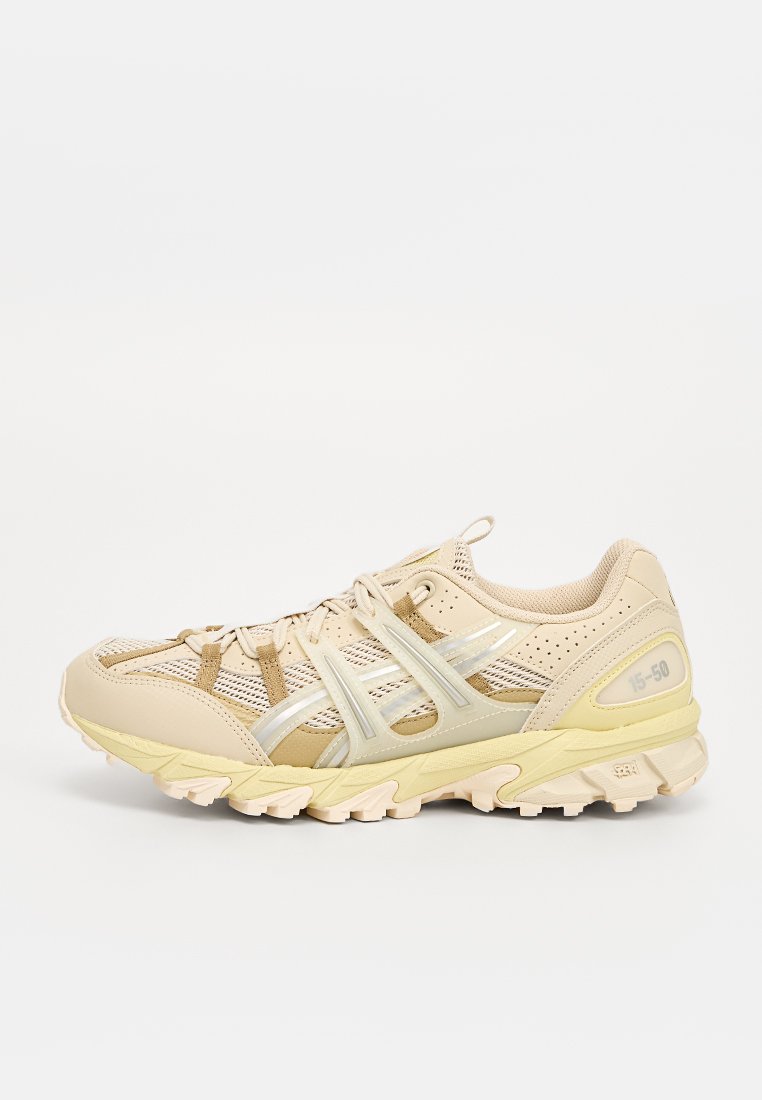 Beige sportssko med nätpaneler, reflekterande silverdetaljer och texturerad sula. Har ett snörningssystem och dämpning för stöd.