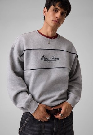 Giovane uomo che indossa una felpa Guess Jeans grigio chiaro con scritta nera, jeans scuri, anelli d'argento e una piccola collana con ciondolo su uno sfondo semplice.