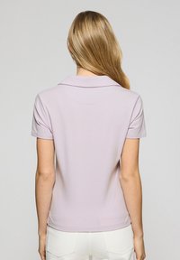 Jasnolavendowy, krótko rękawowy polo z klasycznym kołnierzykiem, gładką teksturą i dopasowanym krojem, noszony z białymi spodniami, widok z tyłu.