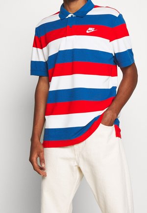 Polo rayé rouge, blanc et bleu avec un col et un petit logo Nike sur la poitrine, associé à un pantalon de couleur claire.