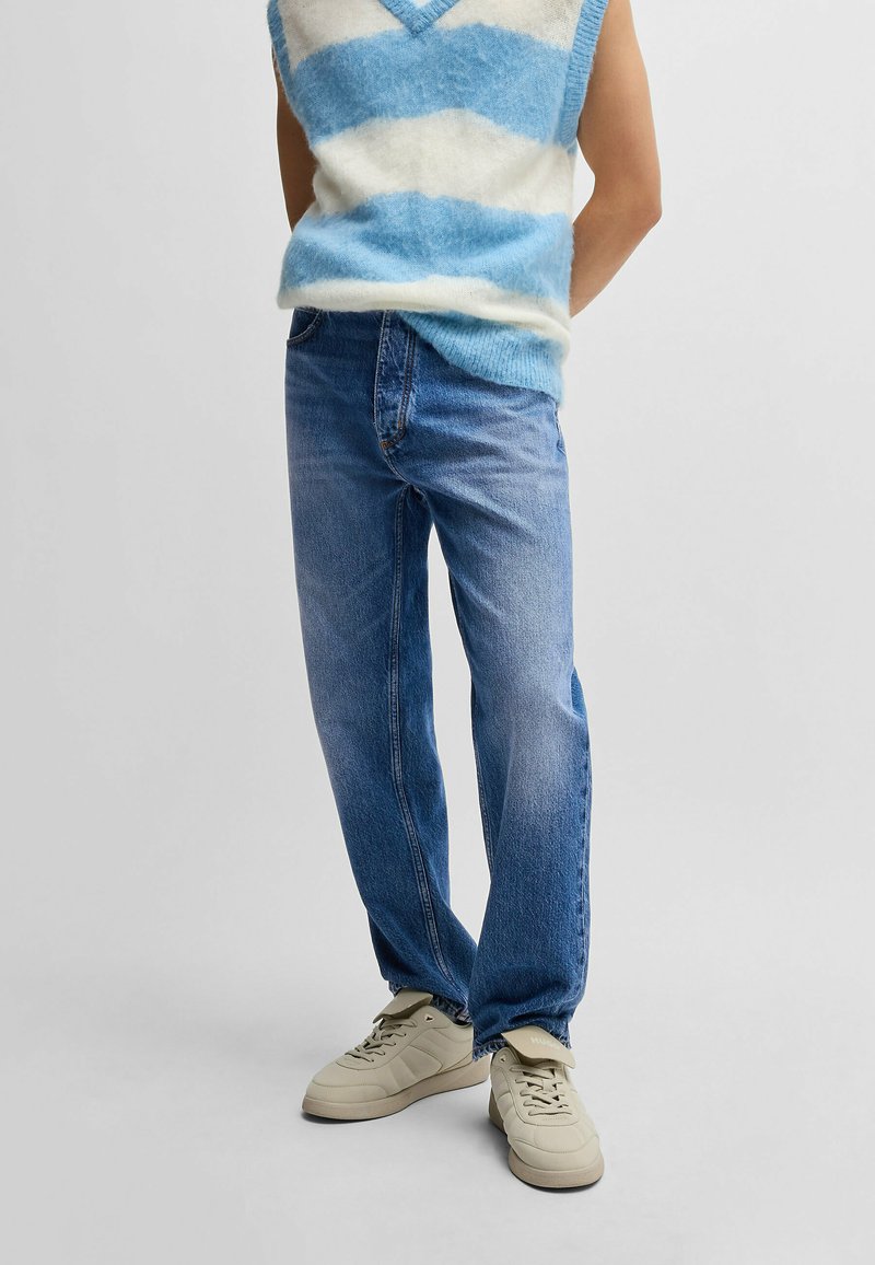 Top sin mangas de punto a rayas azul y blanca combinado con jeans rectos de un tono azul claro y zapatillas beige con un sutil branding y acentos tonales.