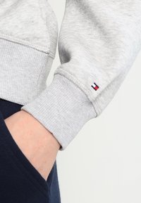 Grijze sweatshirt met geribbelde mouwen, voorzien van een klein logo met rode en blauwe accenten nabij de pols. Zichtbare textuur van de stof.