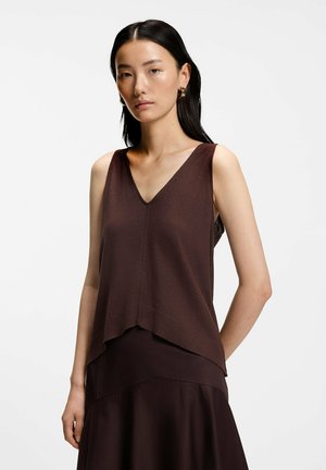 Jeune femme aux longs cheveux noirs porte un haut marron sans manches à col en V et une jupe foncée, faisant face à un fond clair uni.