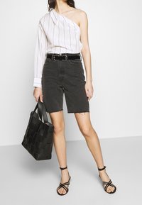 Camisa blanca a rayas con un hombro, pantalones cortos de mezclilla negra, bolso de mano tejido negro y sandalias negras con tiras. Conjunto informal con texturas contrastantes.