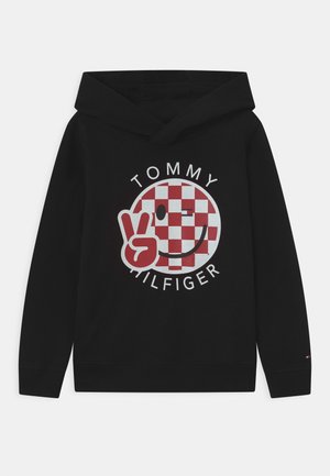 Sort hættetrøje med en cirkulær rød og hvid ternet grafik med et smilende ansigt og fredstegn, mærket "TOMMY HILFIGER" i hvid tekst.