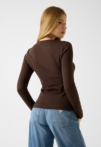 Brązowa bluzka z długim rękawem wykonana z elastycznego materiału, dopasowany krój, okrągły dekolt. Zestawiona z jasnoniebieskimi, wysokim stanem dżinsami z tylną metką z logo.