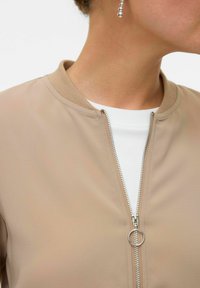 Bež bomber jakna s glatkom teksturom, rebrastim ovratnikom i detaljima srebrnog zatvarača. Nosi se preko bijele majice.