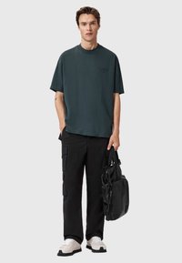 Ciemnozielona bawełniana koszulka z okrągłym dekoltem, zestawiona z czarnymi spodniami cargo. Model trzyma czarną skórzaną torbę; nosi białe sneakersy z czarnymi podeszwami.