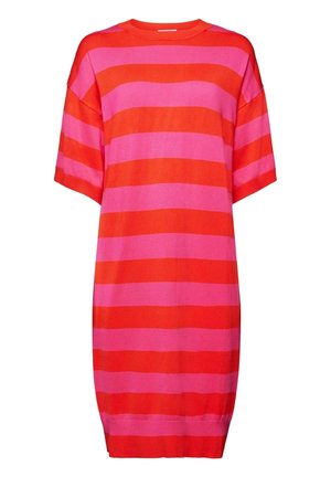 Vrouw die een rood en roze gestreept T-shirt met korte mouwen draagt, met een brede witte riem en een kettinghalsketting tegen een lichtblauwe achtergrond.