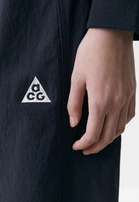 Črne hlače iz blaga z trikotnim belim vezenim logotipom na strani. Blago ima gladko teksturo, ki poudarja čiste šive.