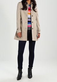 Nainen pukeutuneena vaaleanbeigeen trench-coattiin, värikkääseen raitapaitaan, tummiin slim fit -farkkuihin sekä mustiin nilkkabootseihin valkoista taustaa vasten.