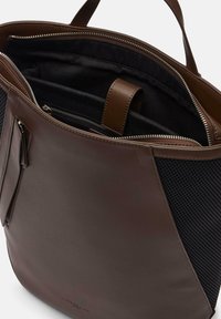 Sac fourre-tout en cuir marron avec un panneau latéral en maillage noir texturé. Dispose d'une fermeture éclair, de compartiments intérieurs et d'une doublure en tissu contrasté.