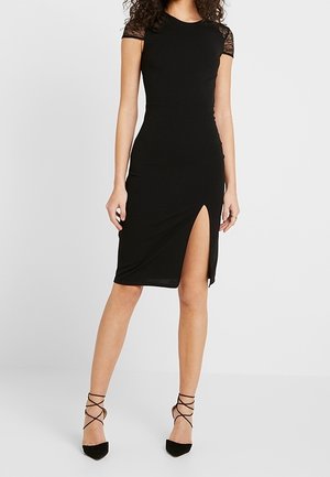 Femme portant une robe noire cintrée longueur genou avec des manches courtes et une fente haute sur le côté gauche, associée à des talons noirs à lanières.