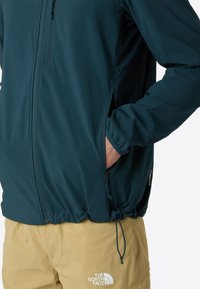 Windbreaker jacket in dark teal, featuring zippered pockets, an adjustable hem, and logo detailing on sleeve. Smooth, lightweight fabric.