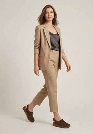 Kobieta idąca pewnym krokiem, ubrana w beżowy garnitur, czarny satynowy top oraz brązowe perforowane buty typu slingback na neutralnym tle.