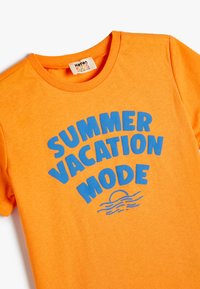Orange bomulds T-shirt med rund halsudskæring. Blå tekst siger "SOMMERFERIE MÅDE" med et simpelt sol- og bølgesymbol nedenfor.