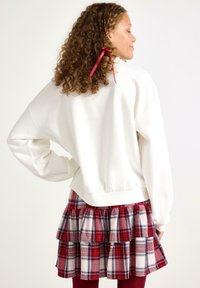 Hvid sweatshirt med afslappet pasform, parret med en rød og ternet lagdelt nederdel med blødt lag. Håret pyntet med et rødt bånd.