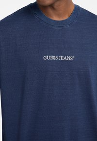 Laisvalaikio tamsiai mėlynas medvilninis marškinėliai su apykakle, kuriuos puošia balta siuvinėta "GUESS JEANS" logotipas ant krūtinės. Minkšta tekstūra, laisvas siluetas.