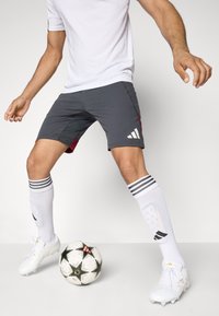 Šedé atletické šortky s marhuľovými akcentami a bielym logom Adidas, spárované s bielymi ponožkami so čiernymi pruhmi a bielymi kopačkami. Vedľa leží futbalová lopta.