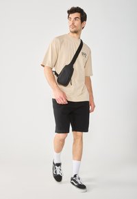 Maglietta oversize beige con stampa floreale, pantaloni corti neri e borsa a tracolla minimalista nera, abbinati a sneaker in bianco e nero e calzini bianchi.