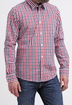 Mann trägt ein rot-weiß-blau kariertes langärmliges Hemd und blaue Jeans, steht vor einem schlichten weißen Hintergrund.