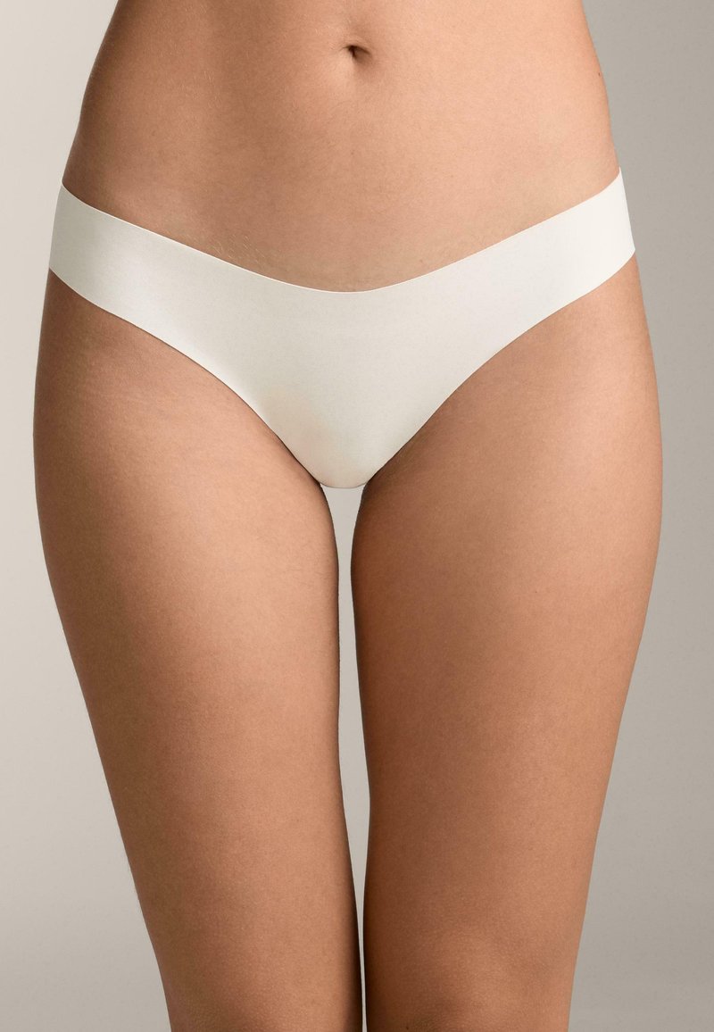 Partie inférieure du torse d'une personne portant des sous-vêtements beiges clairs sans couture sur un fond neutre.