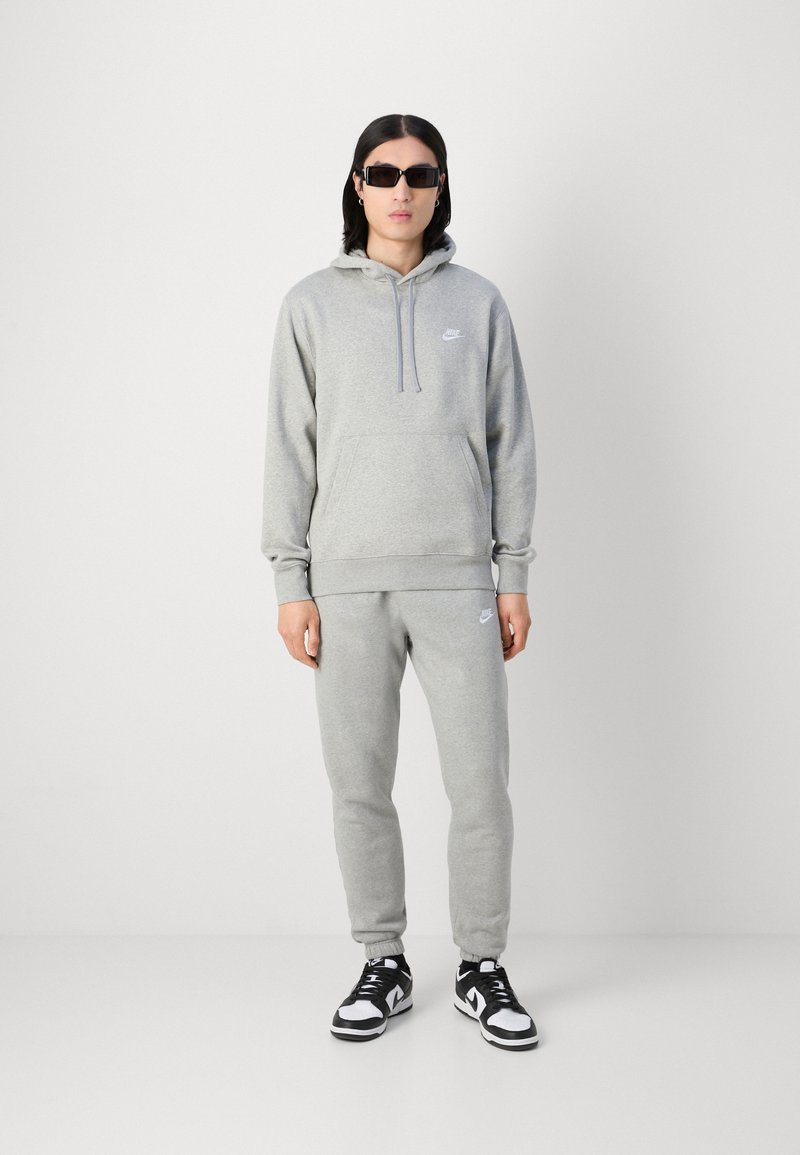 Grau Sweatshirt- und Sweatpants-Set aus weichem Material, mit einer Vorderstasche, Kapuze mit Kordelzug und kleinem Nike-Logo. Kombiniert mit schwarzen Turnschuhen.