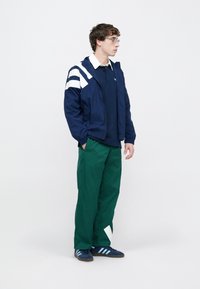 Tengerészkék széldzseki fehér csíkokkal, tengerészkék póló, zöld nadrág, és tengerészkék tornacipő világoskék csíkokkal. Egyszerű, funkcionális kialakítás.