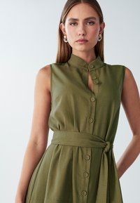 Bežinė suknelė be rankovių su mandarinio tipo apykakle, mygtukų užsegimu priekyje, dirželiu juosmens srityje ir lygiu paviršiumi. Paprastas dizainas su laisvu siluetu.