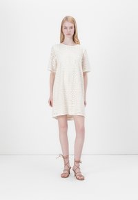 Abito in pizzo di colore avorio con scollo rotondo e maniche corte, caratterizzato da motivi floreali e una vestibilità ampia. Tacchi a strappo marroni completano l'outfit.