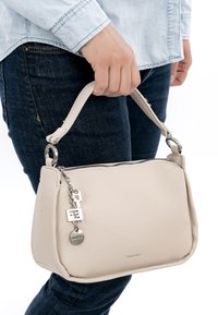 Persoon in denim die een beige gestructureerde handtas vasthoudt met een zilveren ritssluiting en decoratieve bedels, tegen een effen witte achtergrond.