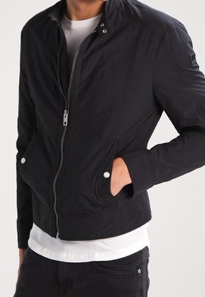 Mann trägt eine schwarze Jacke mit Reißverschluss und Pattentaschen über einem weißen Hemd, mit einer Hand in der Tasche und schwarzen Hosen.