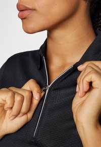 Črna zip-up jopica z medenčastim vzorcem, s srebrnim zadrgo in ovratnikom. Roke držijo zadrgo in jo rahlo vlečejo navzdol.