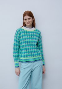 Gestrickter Pullover mit einem blau-grünen Muster, rundem Ausschnitt und gerippten Bündchen. Kombiniert mit hellblauen Cordhosen.