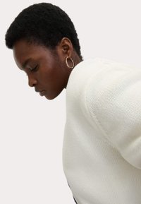 Maglione bianco lavorato a maglia con tessuto testurizzato e scollatura rotonda, abbinato a un semplice orecchino a cerchio in oro. La luce soffusa mette in evidenza i dettagli.