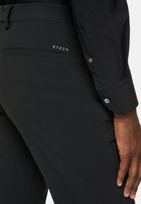 Svarta byxor med en slät yta, med en bakficka och en liten vit logotyp. Midjan har snörning och knappdetaljer.