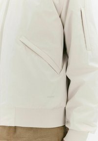 Licht beige bomberjack met een ritssluiting, zijzak, ribgebreide boorden en een gestructureerde zoom. Gemaakt van gladde, waterafstotende stof.