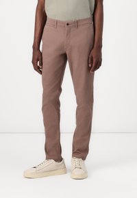 Pantalones de algodón marrón claro con corte recto, presentan un cierre de botón en la parte frontal, cinco bolsillos y pasadores para cinturón. Combinados con zapatillas beige.