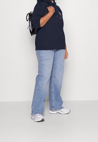 Felpa blu navy con cappuccio e cordino, jeans dritti azzurri chiari e sneakers bianche. L'individuo porta uno zaino nero piccolo.