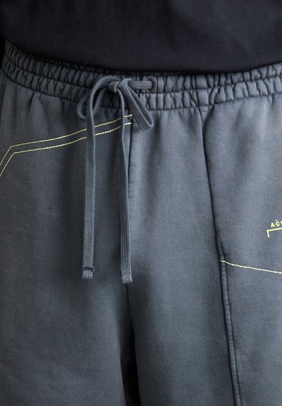 Graue Trainingshosen mit einem elastischen Bund und Kordelzug. Enthält gelbe Nähte in einem geometrischen Muster. Weiche, baumwollähnliche Textur.