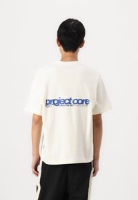 Elfenbensfarvet T-shirt med korte ærmer, der har stor blå tekst "project core" og lille logotryk på ryggen, med en afslappet pasform og sideslids.