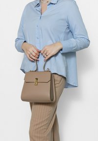 Borsa strutturata beige con chiusura dorata e manici corti, realizzata in pelle liscia, con un design minimalista. Visibili pantaloni a righe.
