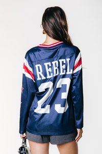 Marineblå langermet genser med hvit "REBEL" tekst og nummer "23" på baksiden. Har røde og hvite stripeakenter på ermene. Glatt tekstur.